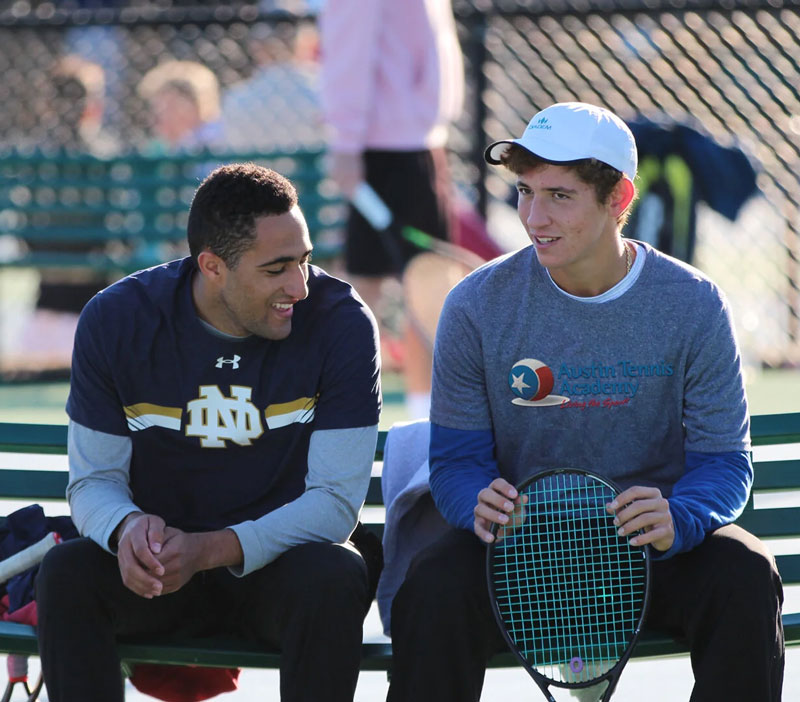 Two young men sit on a bench, smiling and talking, one holding a tennis racket on a sunny day.