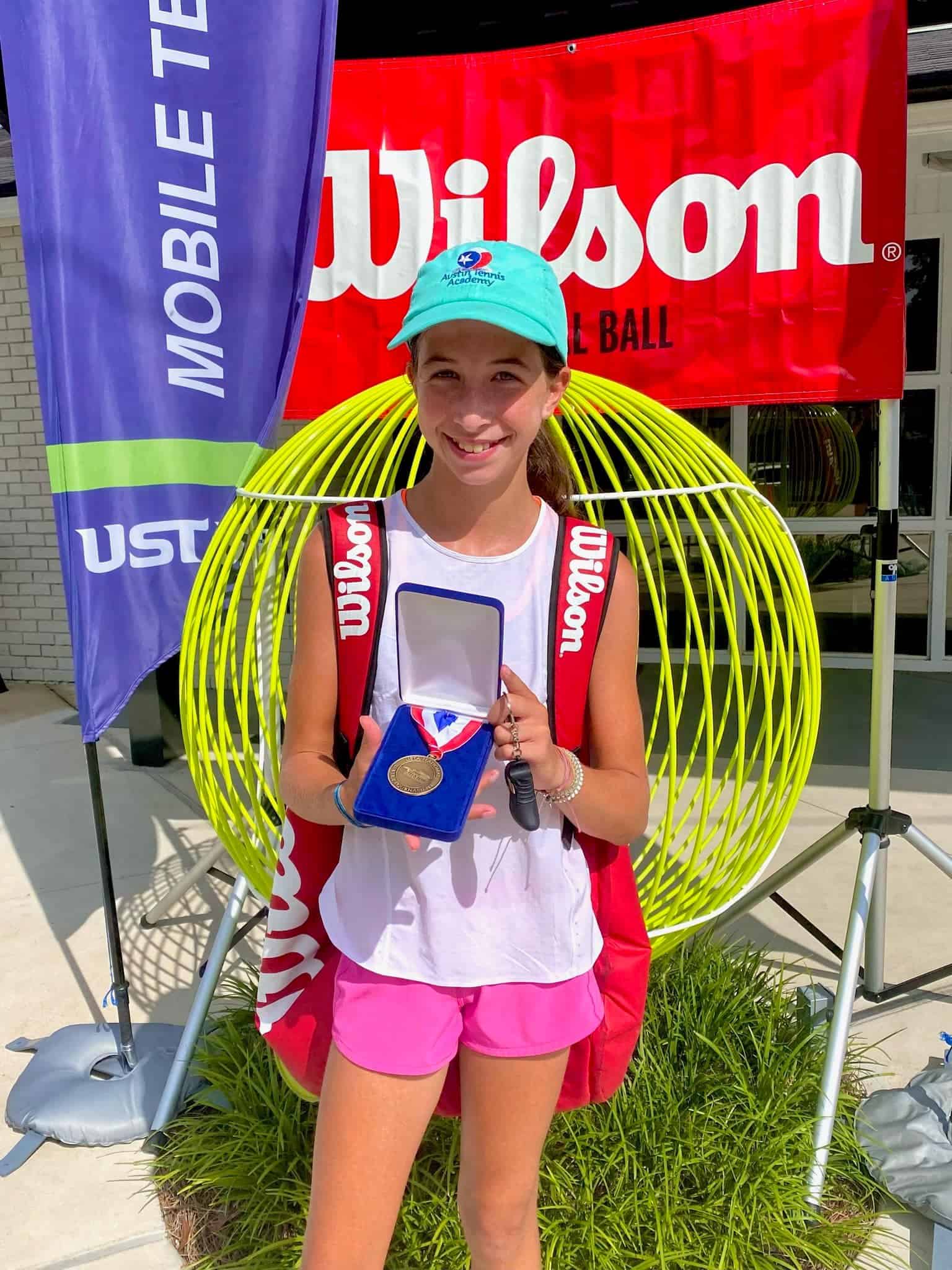 Smiling girl in athletic wear holds a medal in front of Wilson tennis display and event banners outdoors.