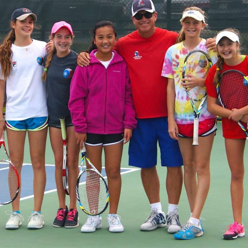 A tennis coach stands with five smiling girls holding rackets on an outdoor tennis court.