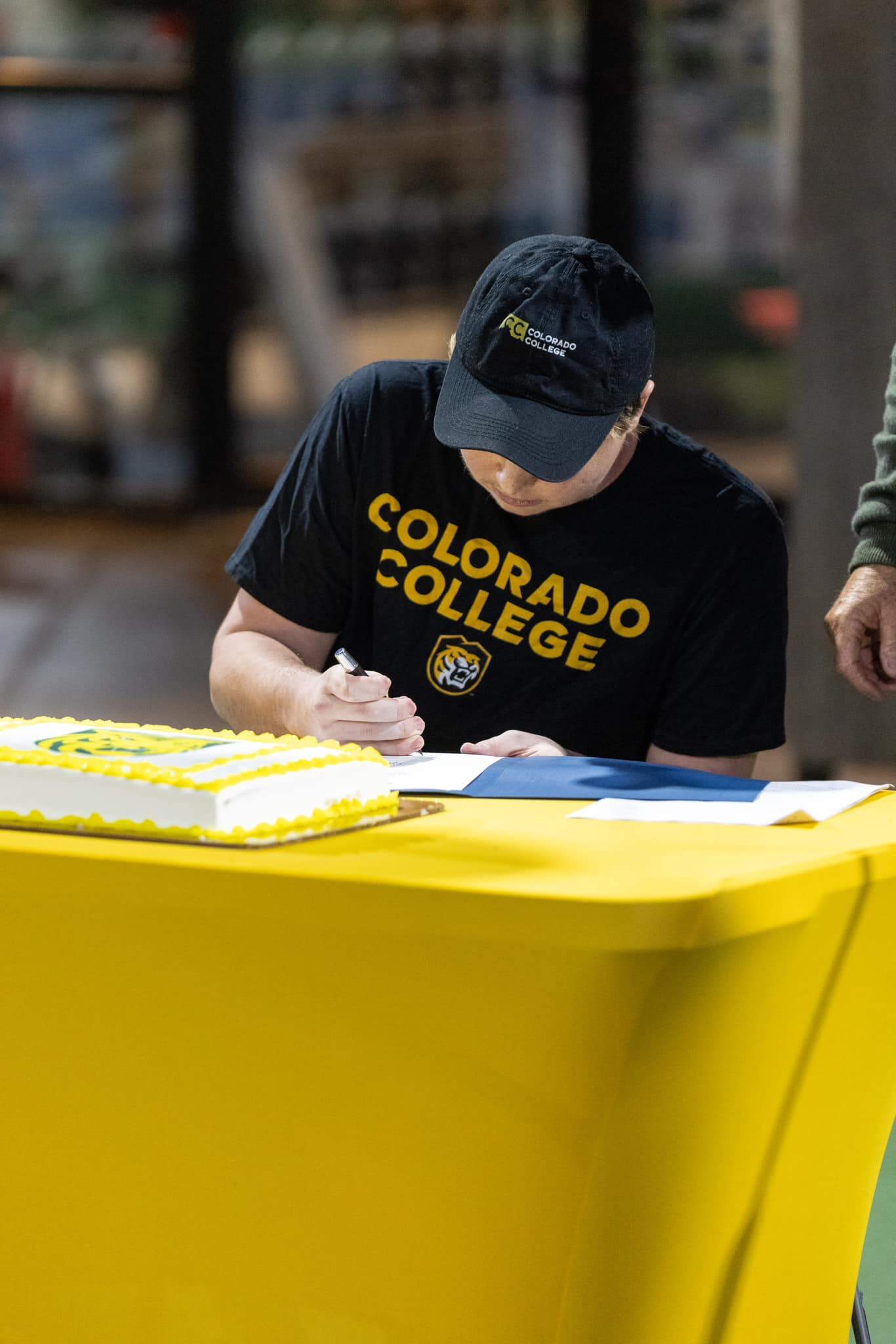 Person in a Colorado College shirt and cap signs a document at a yellow table with a cake nearby.