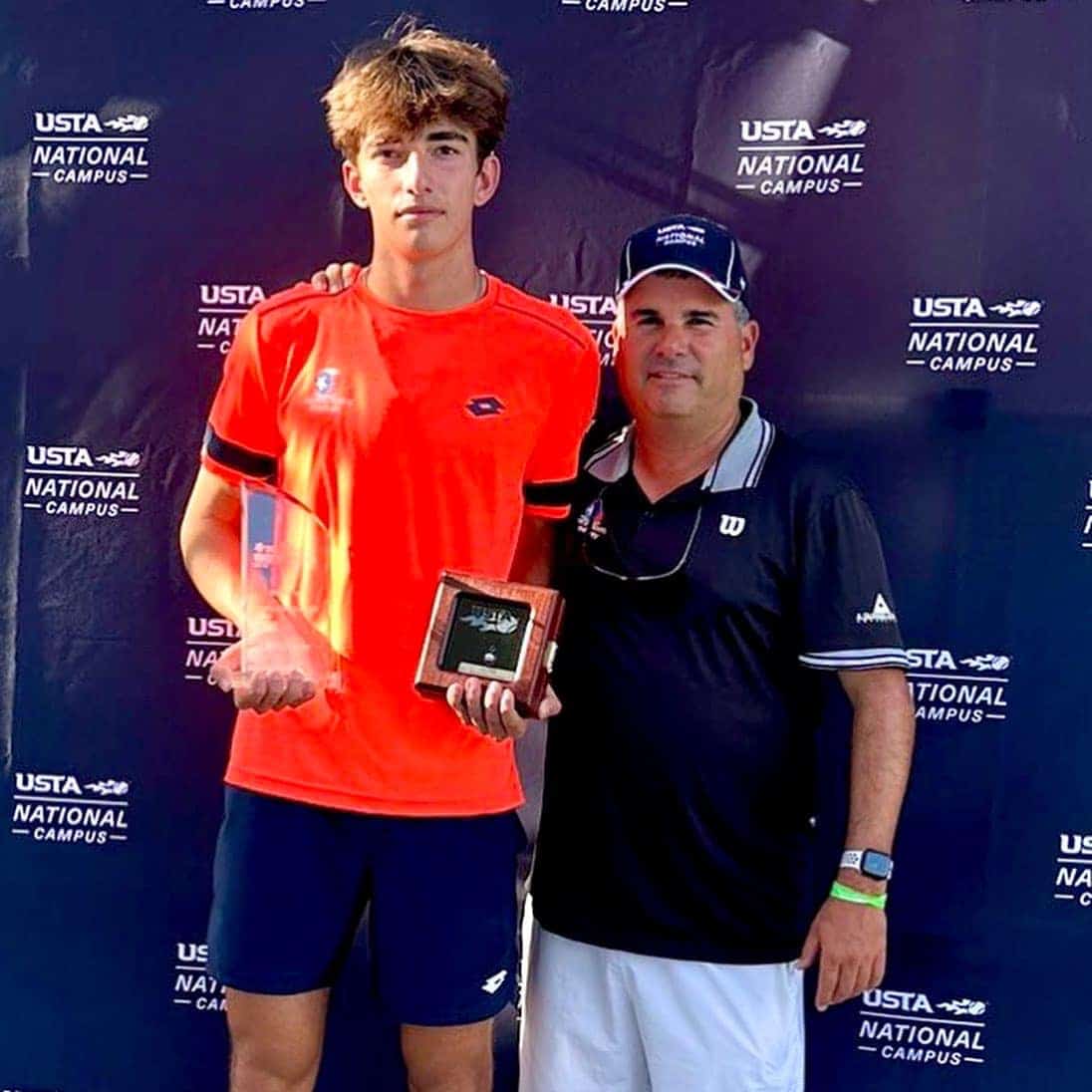 Teen tennis player holding a trophy stands with a man in front of a USTA National Campus backdrop.