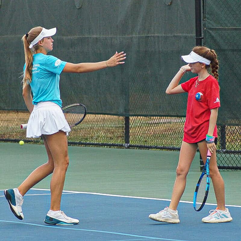 Two girls with tennis rackets talk on a court; one gestures with her arm extended, both wearing visors.