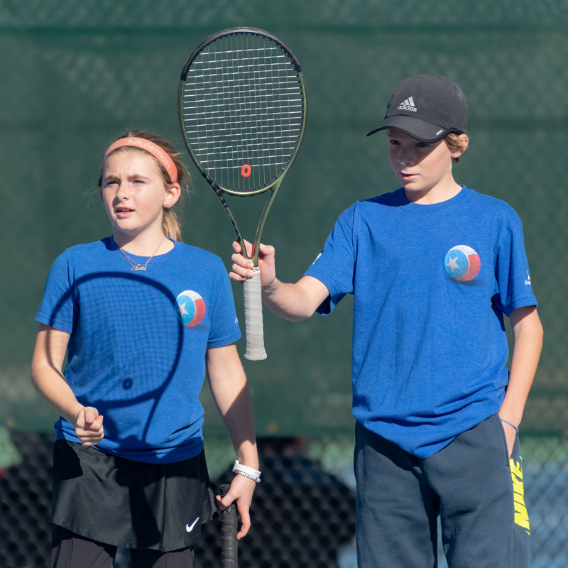 Two kids in blue shirts holding tennis rackets on a court, looking intently toward something off camera.