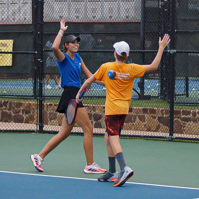 Two people on a tennis court high-five and smile, both wearing sports clothes and holding tennis rackets.