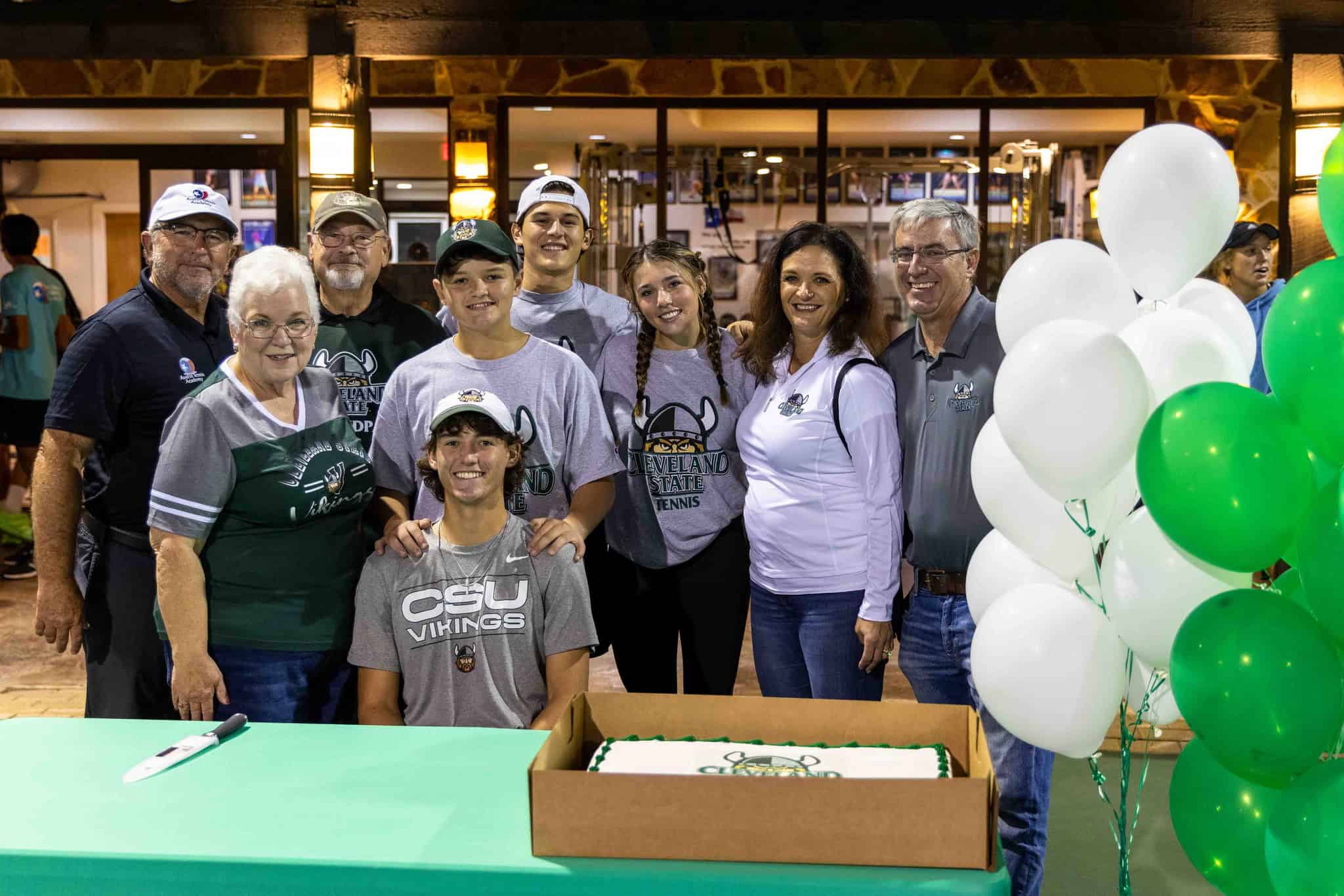 A group of people poses indoors with balloons and a cake, celebrating a young man in a CSU Vikings shirt.