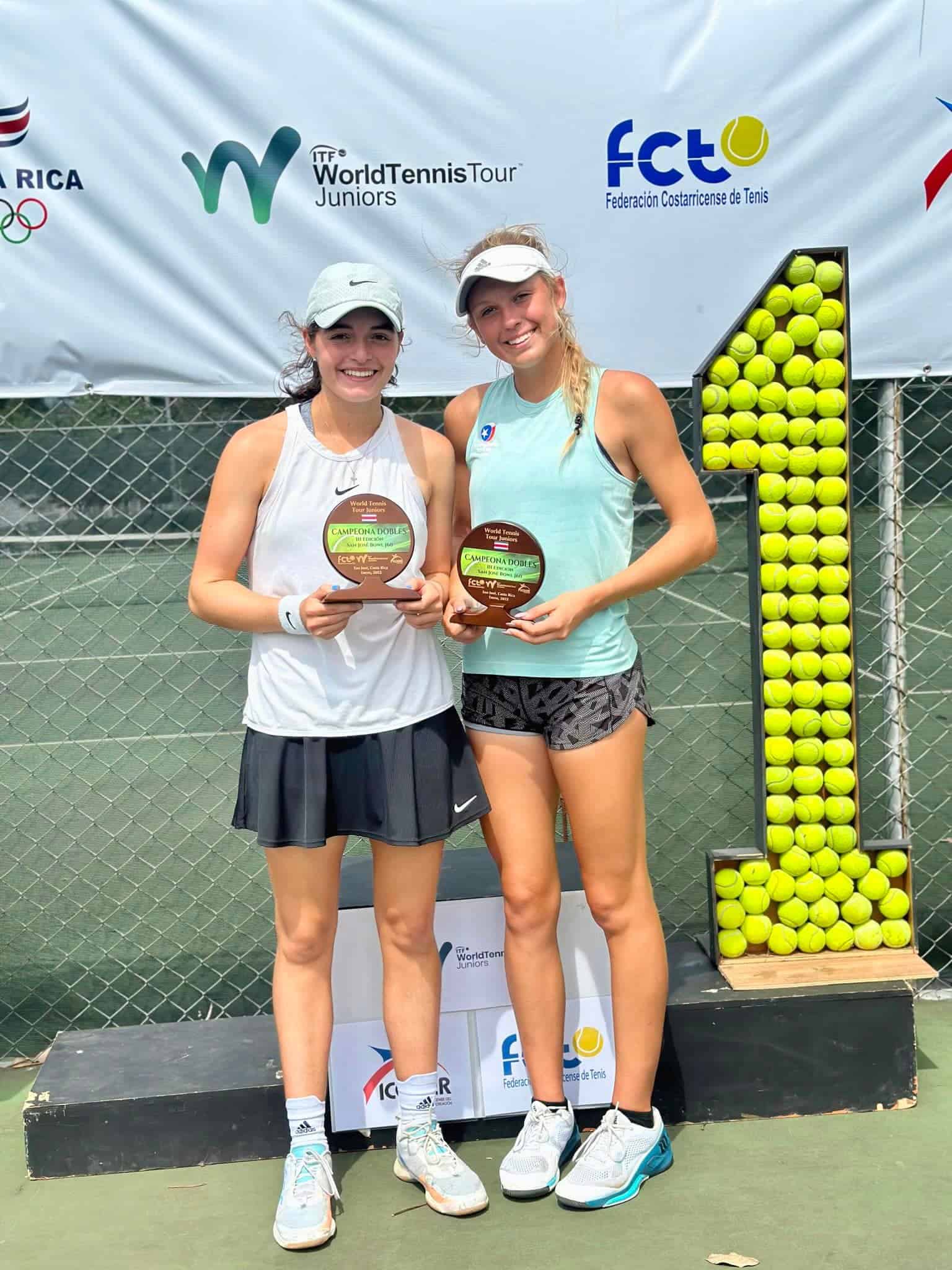 Two young female tennis players holding trophies, standing by a large number 1 made of tennis balls.