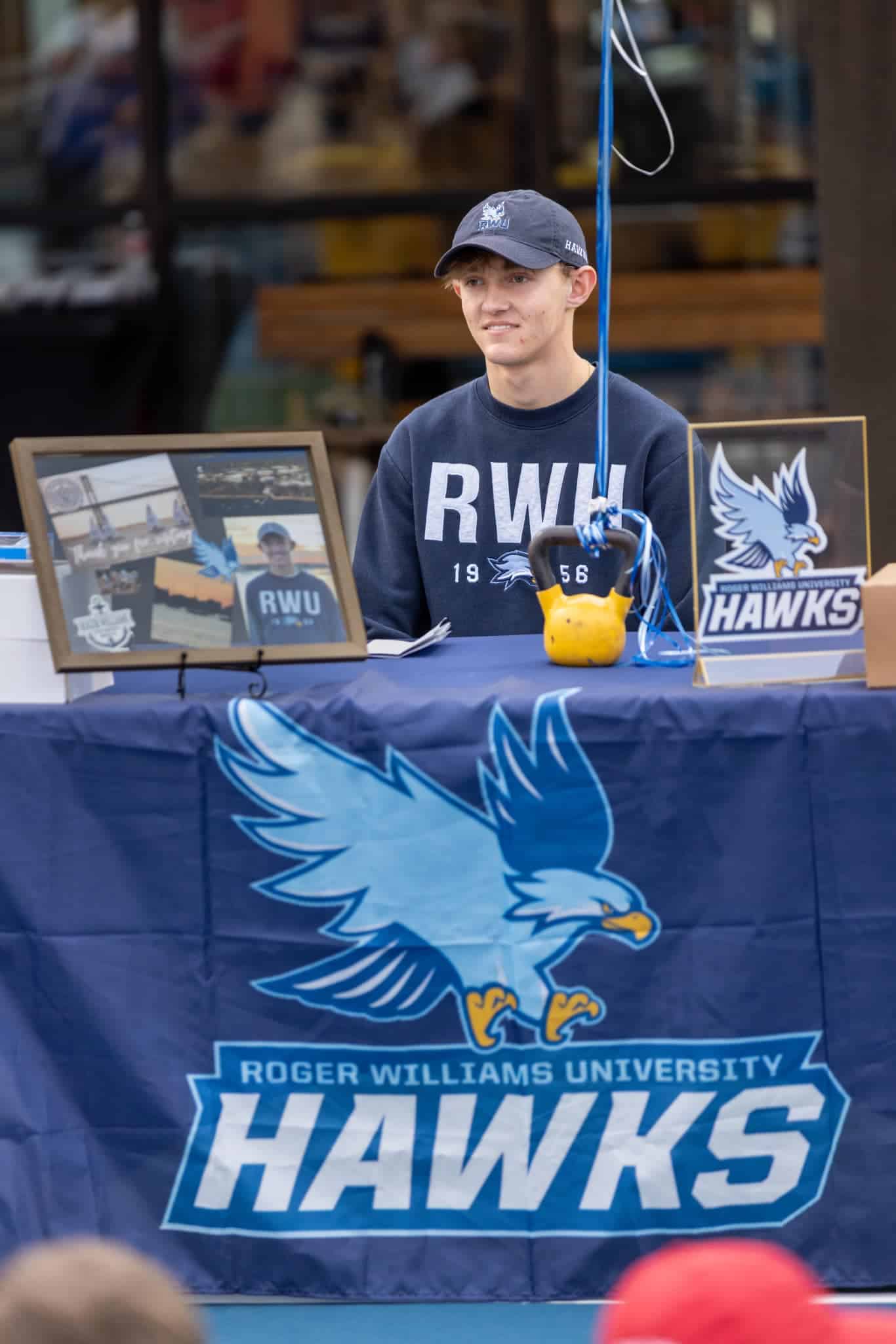 A person sits at a table with Roger Williams University Hawks logos and memorabilia on display.