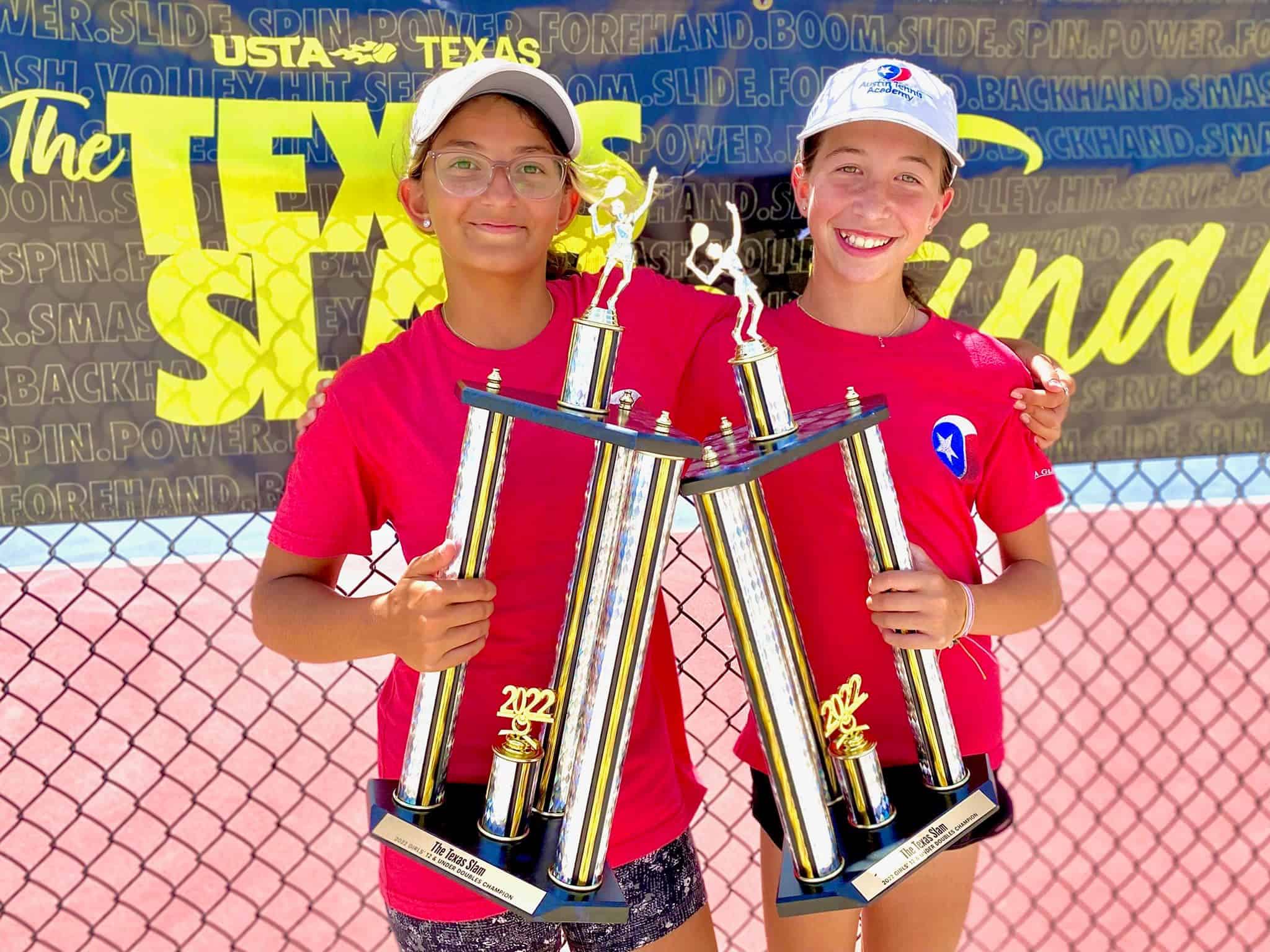 Two smiling girls in red shirts hold tall tennis trophies in front of a Texas tennis tournament banner.