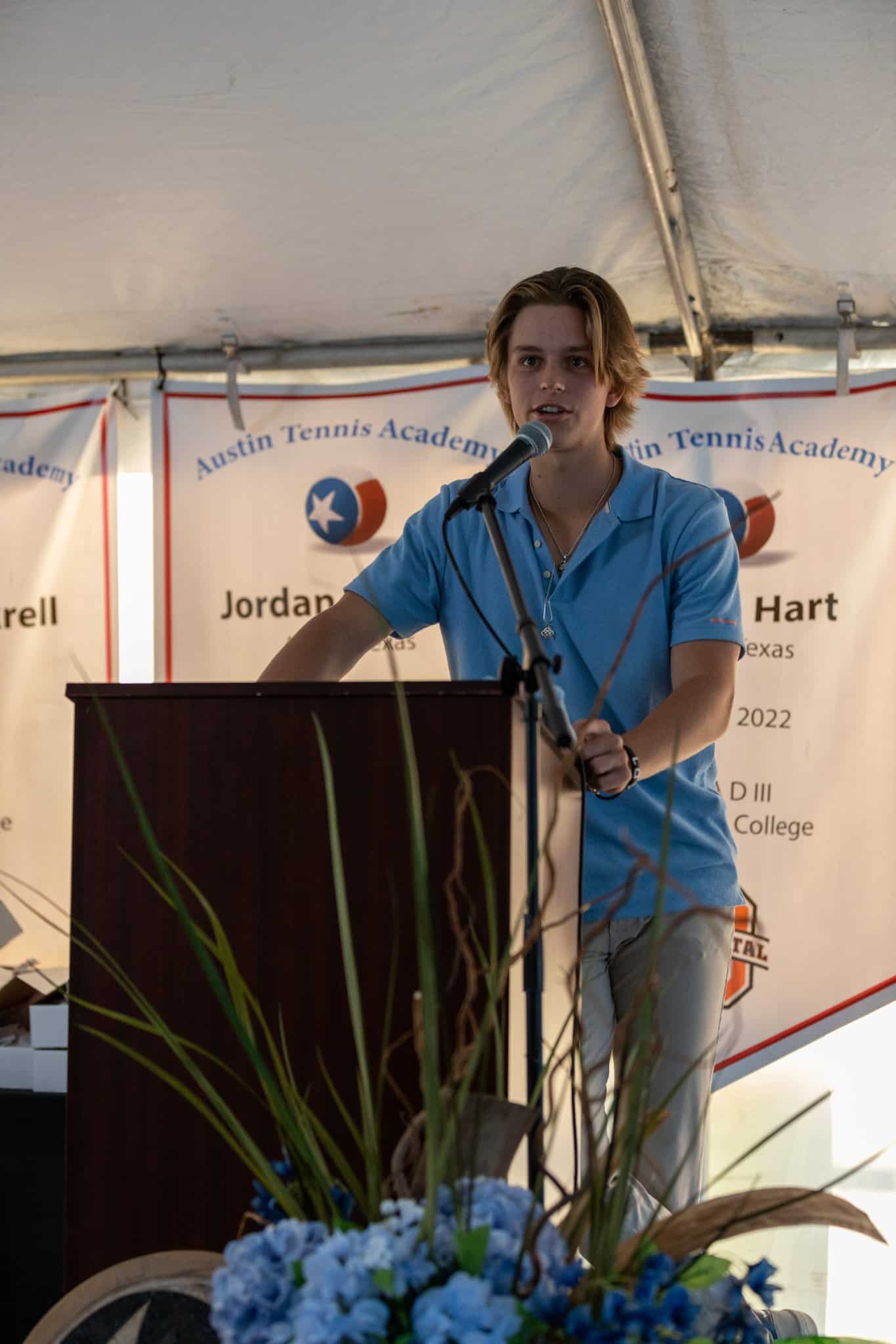 Young man in a light blue shirt speaks at a podium under a tent, with banners in the background.