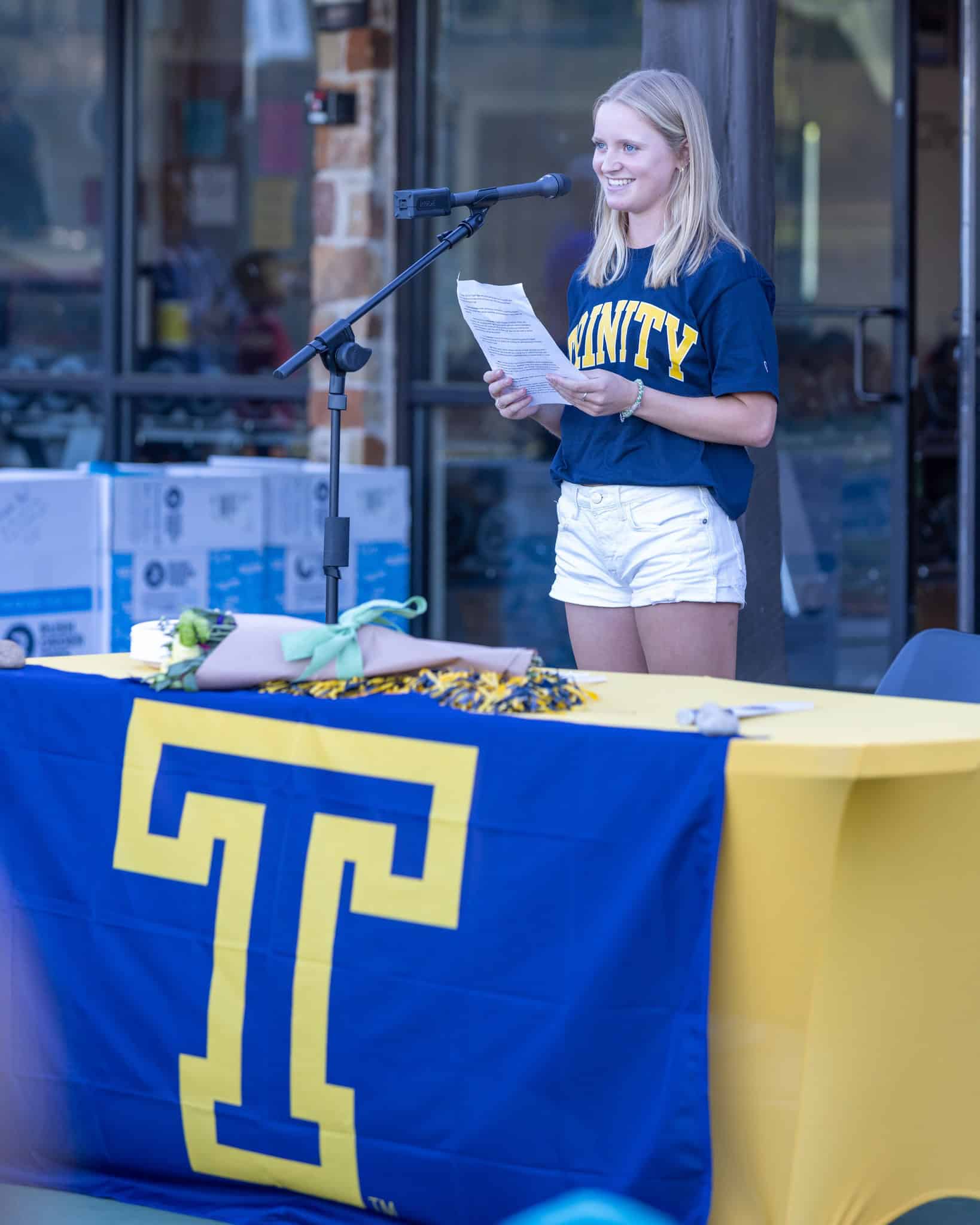 Young woman in a TRINITY shirt speaks at a podium with a large blue and yellow T banner on the table.