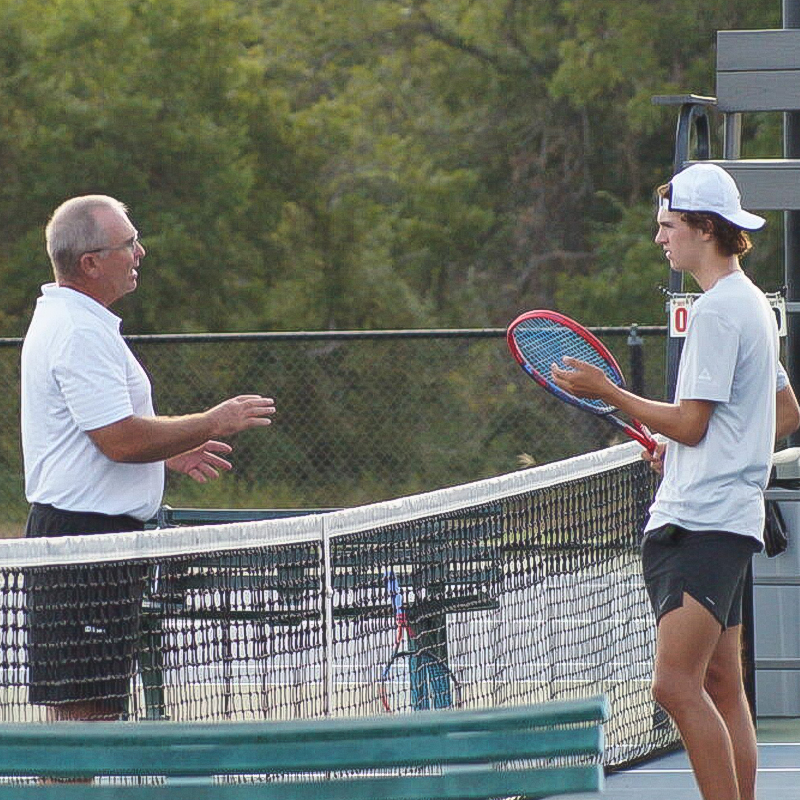 An older man and a young man talk across a tennis net; both hold rackets on an outdoor court.
