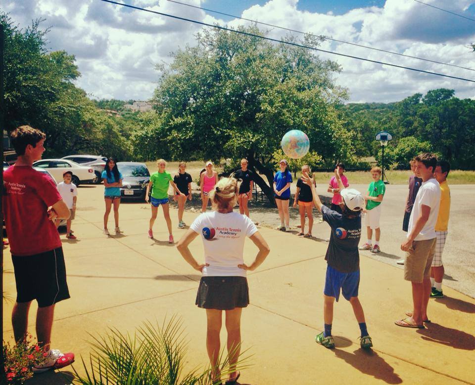 A group of kids and adults stand in a circle outdoors, playing with a beach ball on a sunny day.