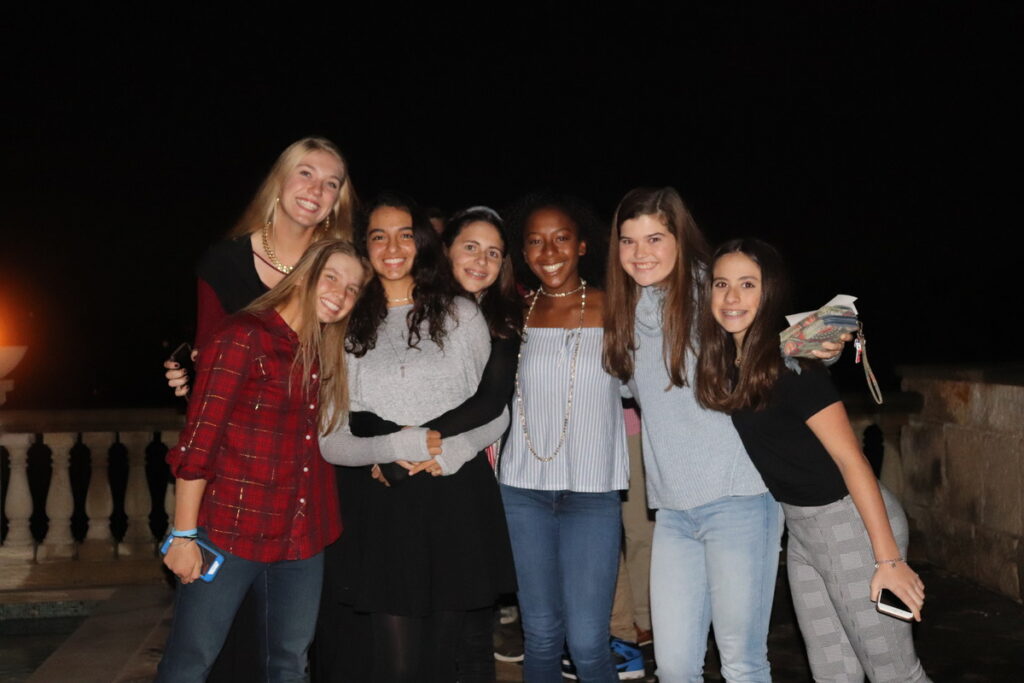 Seven smiling teenage girls pose together outside at night, with arms around each other.