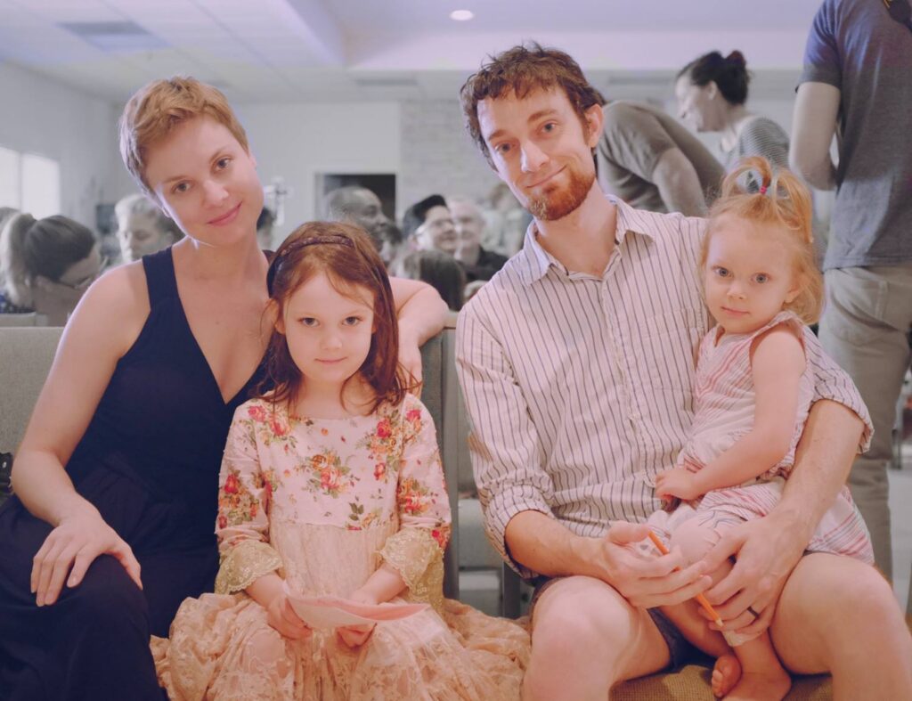 A family of four poses together, seated, with others gathered in the background at an indoor event.