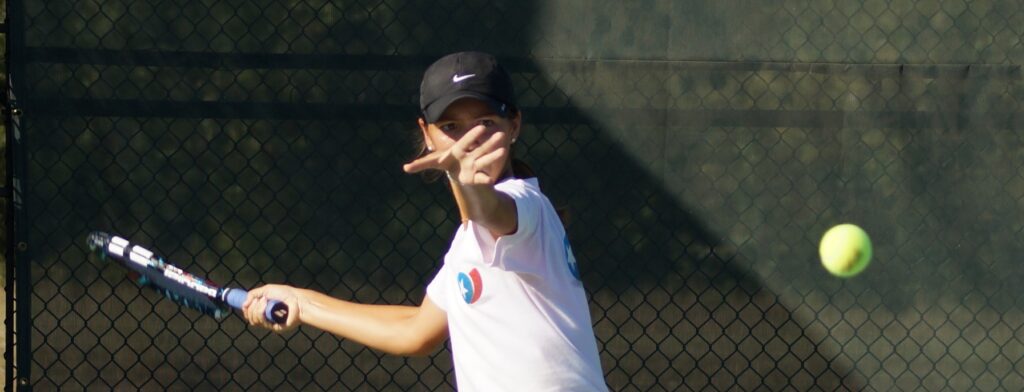 Person in a white shirt and black cap playing tennis, throwing the ball to serve on a court.
