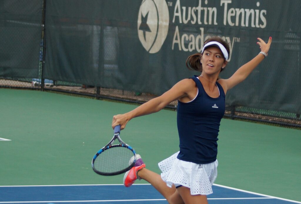 Woman in tennis attire hitting a forehand on an outdoor court, with Austin Tennis Academy sign in background.