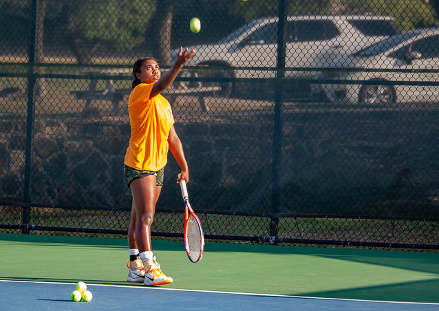 A person in an orange shirt serves a tennis ball on an outdoor court with tennis balls at their feet.