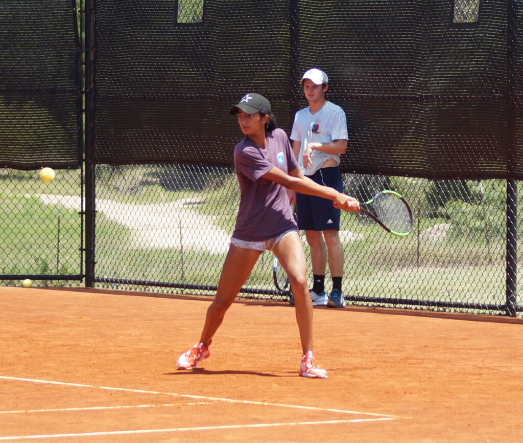 A tennis player prepares to hit a backhand shot on a clay court, with a coach watching in the background.