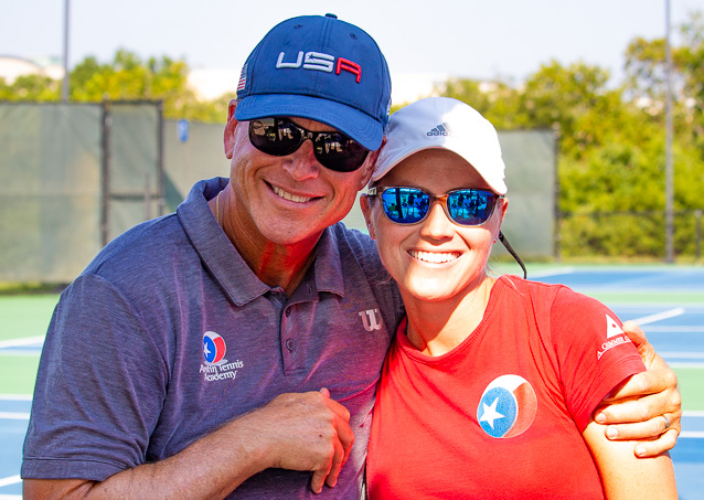 Two smiling people in sportswear and sunglasses pose together on a sunny outdoor tennis court.