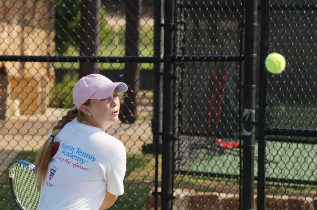A young girl in a pink cap prepares to hit a tennis ball on an outdoor court.