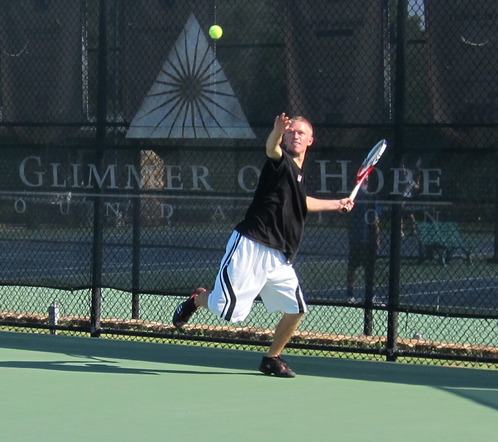 Man in black shirt and white shorts serves a tennis ball on an outdoor court during daylight.
