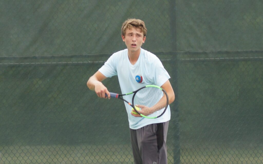 Teen boy in a white T-shirt playing tennis, holding a racket and preparing to hit a ball on an outdoor court.