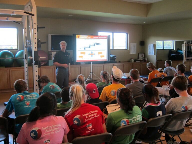 A group of students listens to a man presenting a slideshow in a classroom with gym equipment.