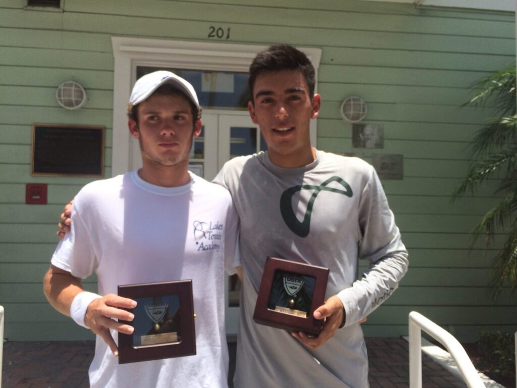 Two young men holding trophies pose together outside a green building with the number 201 above the door.