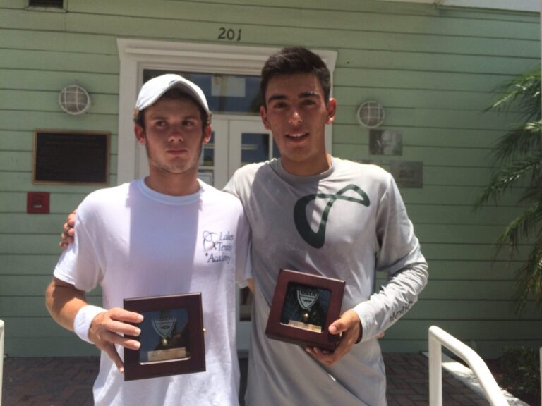 Two young men holding trophies pose together outside a green building with the number 201 above the door.