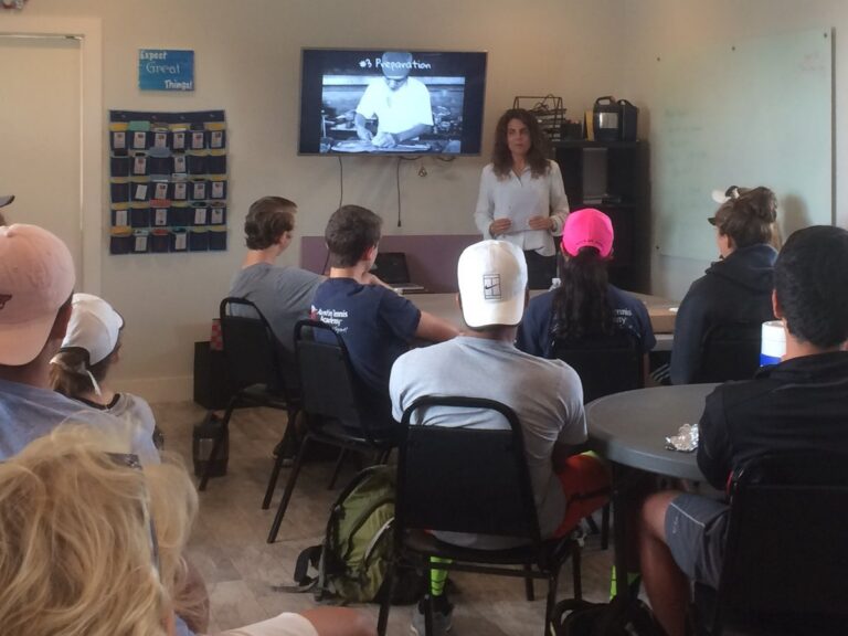 A woman speaks to a group of people seated in a classroom, with a presentation on a screen behind her.