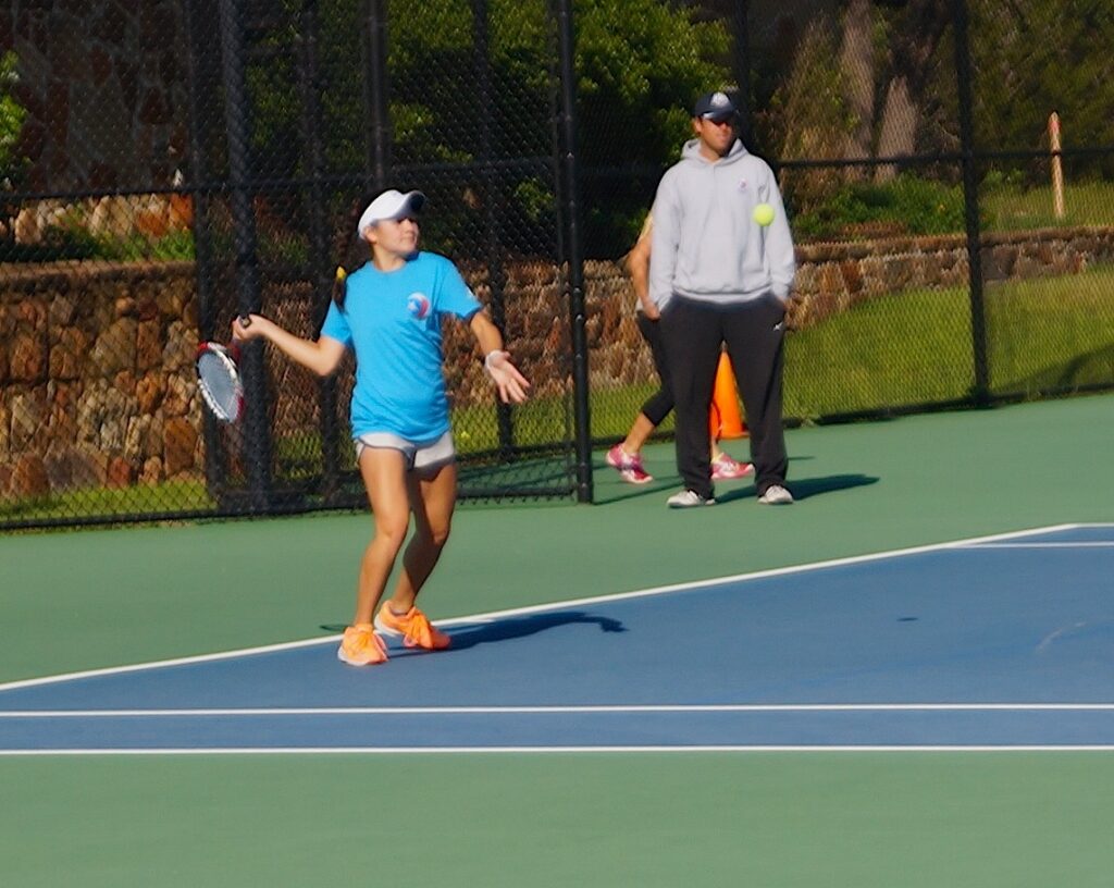 A tennis player in a blue shirt prepares to hit a ball as a coach watches on an outdoor court.