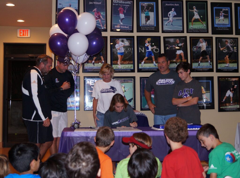 A girl signs a document at a table with balloons, surrounded by adults, as children watch.
