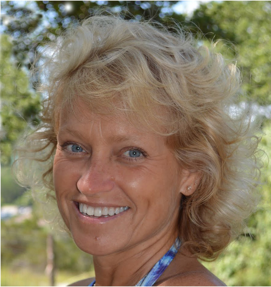 Smiling woman with curly blonde hair, blue eyes, and a blue halter top, outdoors with trees in the background.