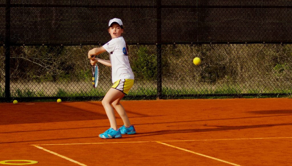 A young tennis player in a white shirt prepares to hit a ball on a clay court.