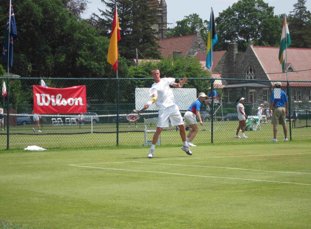 A tennis player in white swings a racket on a grass court during a match, with flags and spectators in the background.