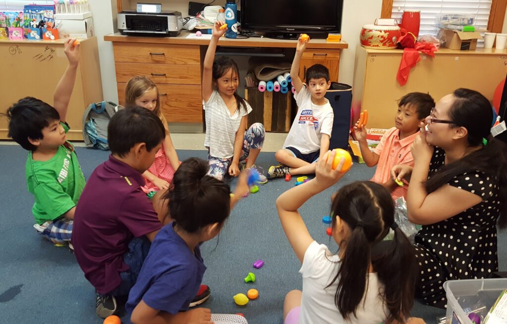 Children sit in a circle on the floor in a classroom, some raising hands and holding colorful toys.