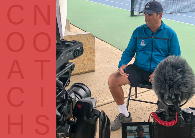 A tennis coach is interviewed courtside, surrounded by cameras and recording equipment.