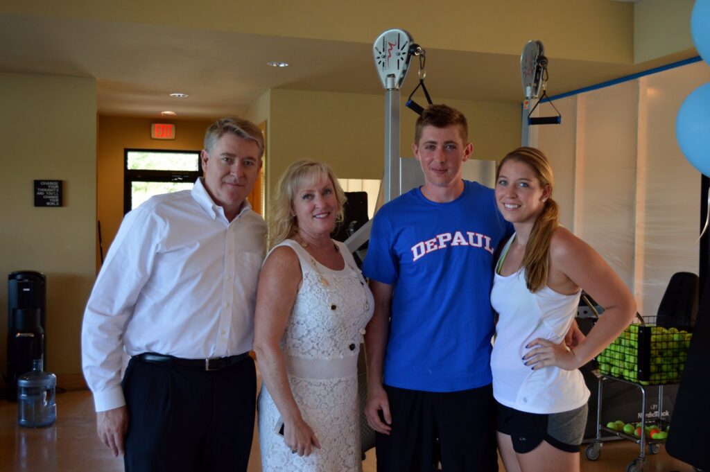 Four people posing indoors, with gym equipment and a cart of tennis balls in the background.