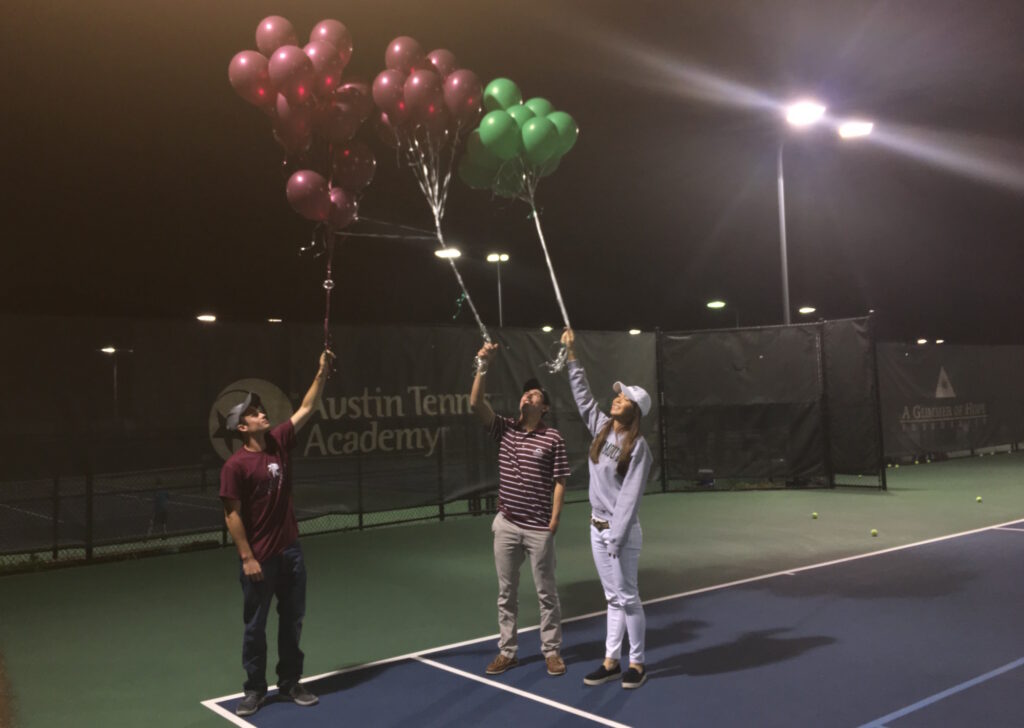 Three people on a tennis court at night, each holding bunches of balloons and reaching them into the air.