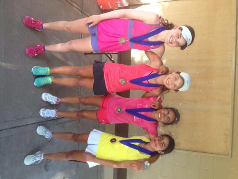 Four girls in athletic wear and medals smile and pose together on a sidewalk near a beige wall.