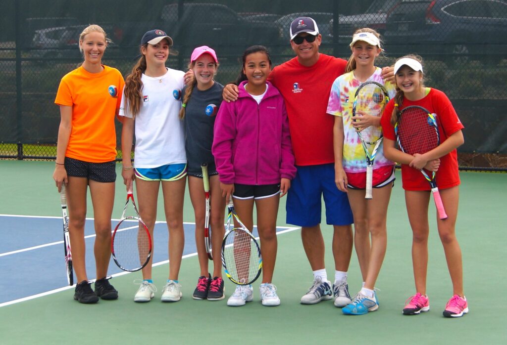 A group of six girls and a coach stand smiling on a tennis court, holding tennis rackets.