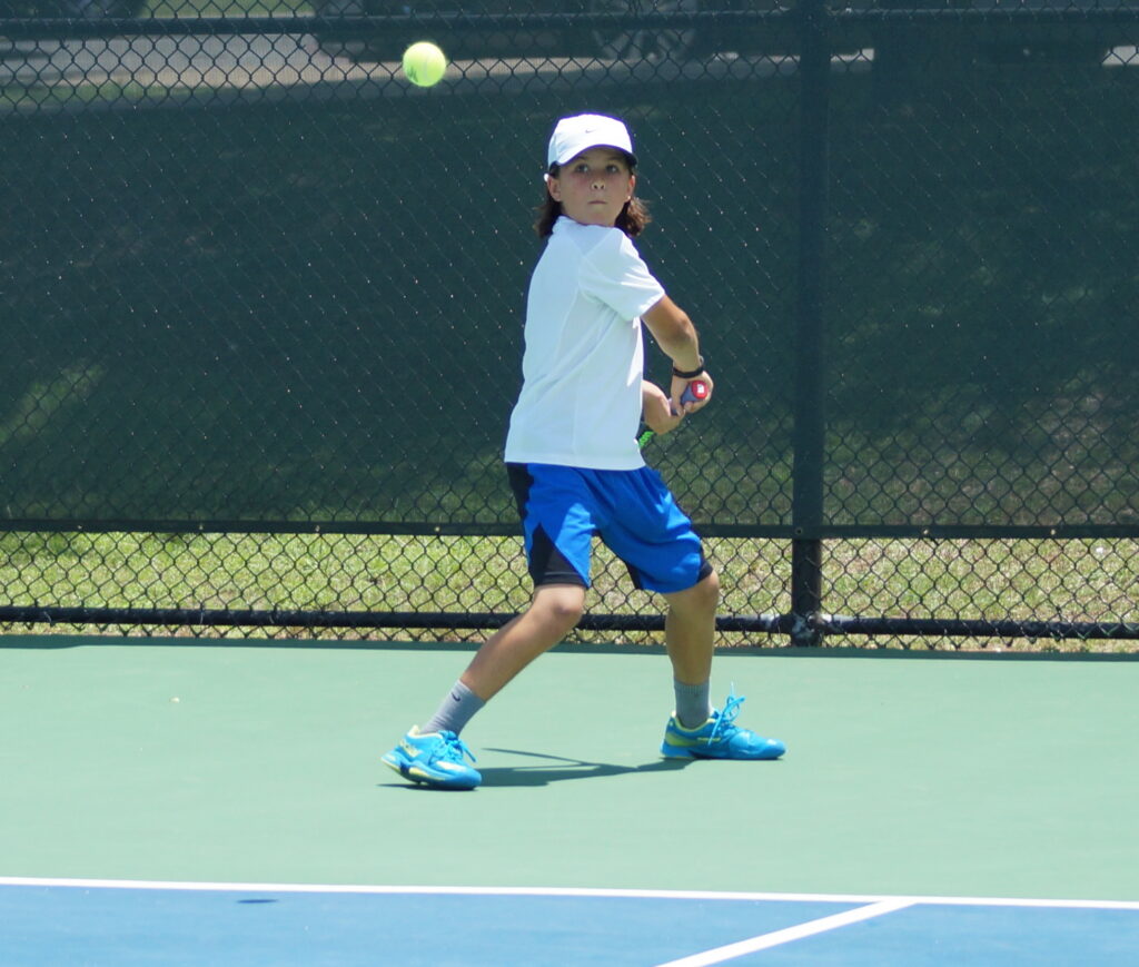 A young boy prepares to hit a tennis ball with a backhand swing on an outdoor court.