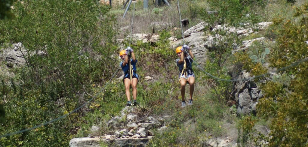 Two people wearing helmets and harnesses ziplining side by side over a rocky, green landscape.