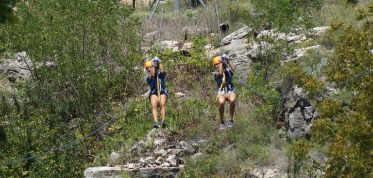 Two people wearing helmets and harnesses ziplining side by side over a rocky, green landscape.