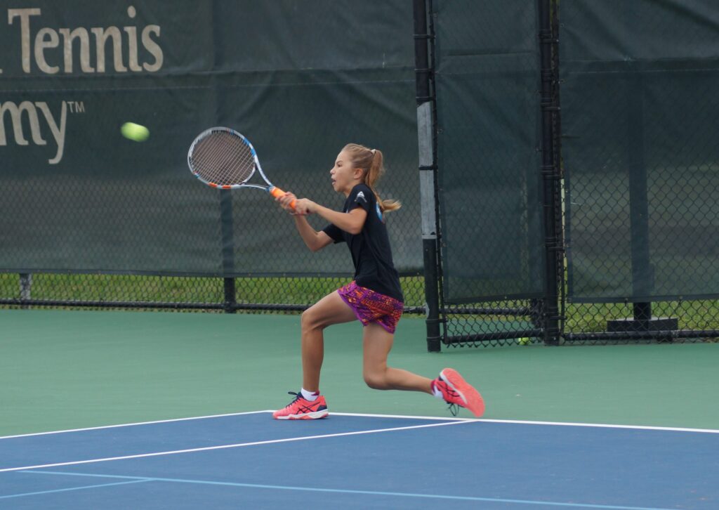 Young girl in sportswear runs to hit a tennis ball with her racket on an outdoor tennis court.