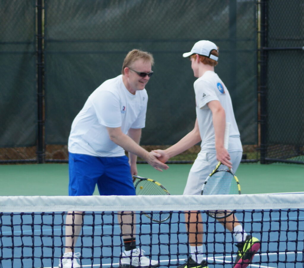 Two tennis players shake hands at the net on an outdoor court, both smiling and holding rackets.