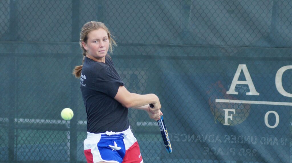 A tennis player in mid-swing watches the ball, wearing a black shirt and red, white, and blue shorts.