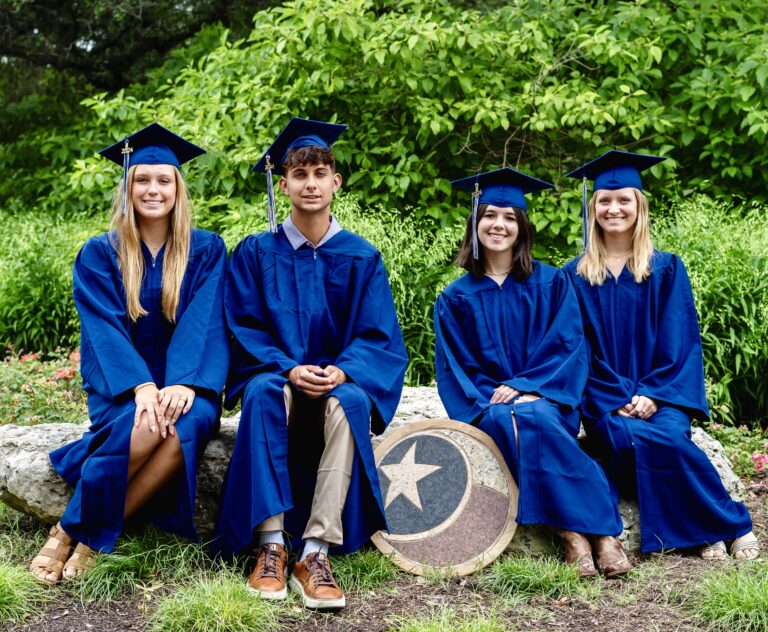 Four graduates in blue caps and gowns sit on a rock outdoors, smiling at the camera.