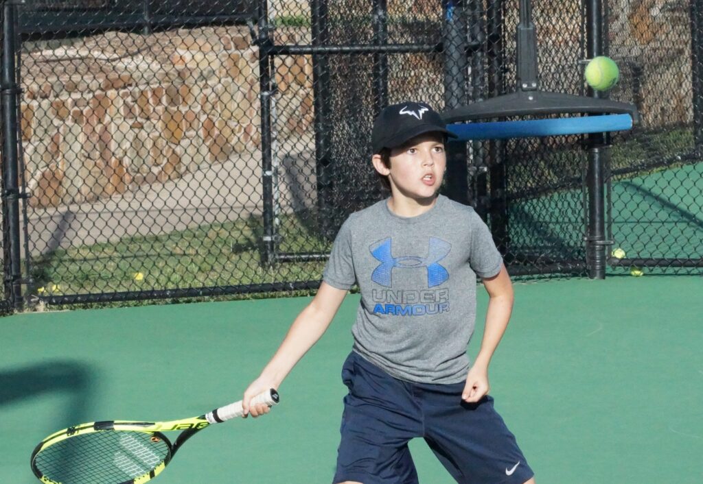A boy in sportswear prepares to hit a tennis ball with a racket on an outdoor court.