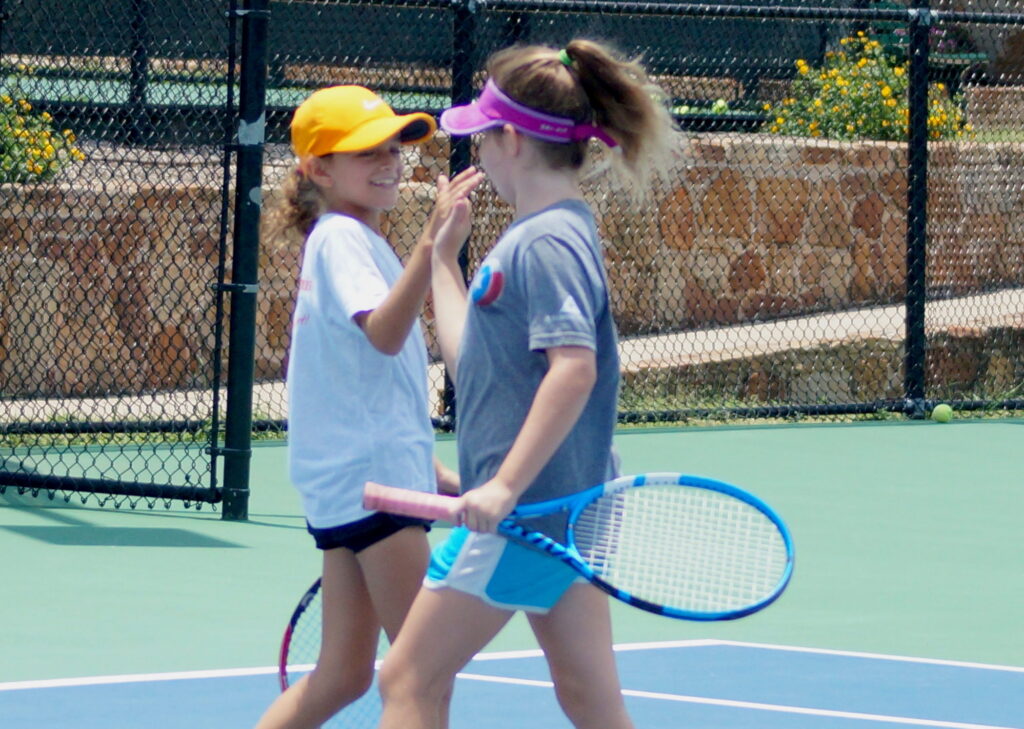 Two young girls on a tennis court, smiling and giving each other a high-five while holding tennis rackets.